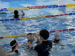 Skywhale coach helping two Kiddie-program children adjust their swim goggles in the pool